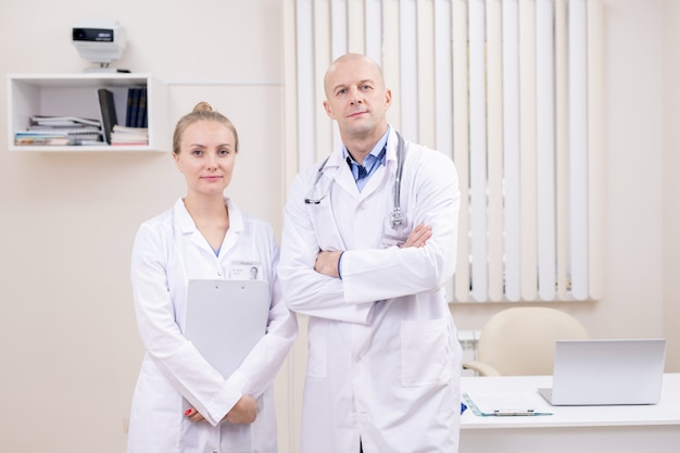 Zwei selbstbewusste Kollegen in medizinischer Uniform, die am Arbeitsplatz in Kliniken nahe beieinander stehen