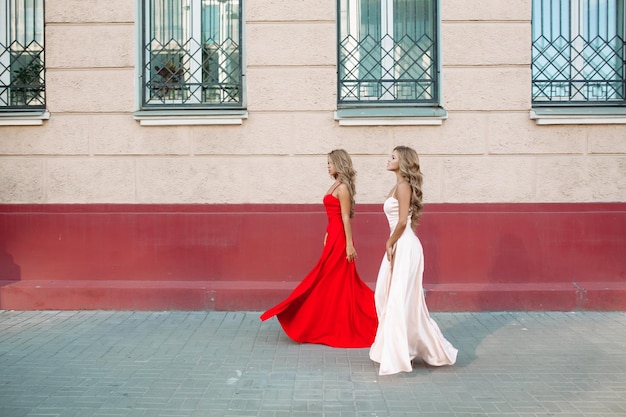 Zwei selbstbewusste und modische Mädchen, die elegante Abendkleider tragen Schlanke, attraktive Frauen mit blonden langen Haaren, die vorwärts gehen und ihre roten und puderlangen Kleider hochsteigen Windiger Effekt