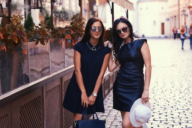 Zwei sexy Brünette in stylischen schwarzen Kleidern mit Sonnenbrille stehen in der Nähe eines Terrassencafés in der Stadt.