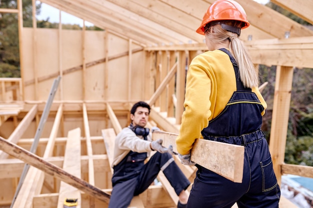 Zwei Spezialisten, die beim Cottage-Bau die Größe des Holzbalkens messen. Immobilienprojekt mit Bauingenieur. Mann und Frau in Uniform helfen sich gegenseitig, entgegenkommend und hilfsbereit