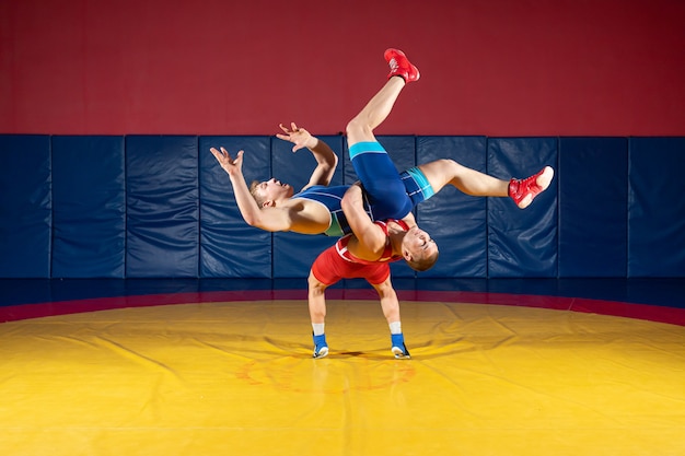 Zwei starke Wrestler in blauen und roten Wrestling-Strumpfhosen machen im Fitnessstudio einen Hüftwurf auf einem gelben Wrestling-Teppich.