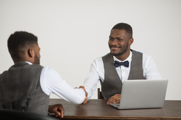 Zwei stilvolle afrikanische Männer in Anzügen sitzen mit einem Laptop an einer weißen Wand am Tisch