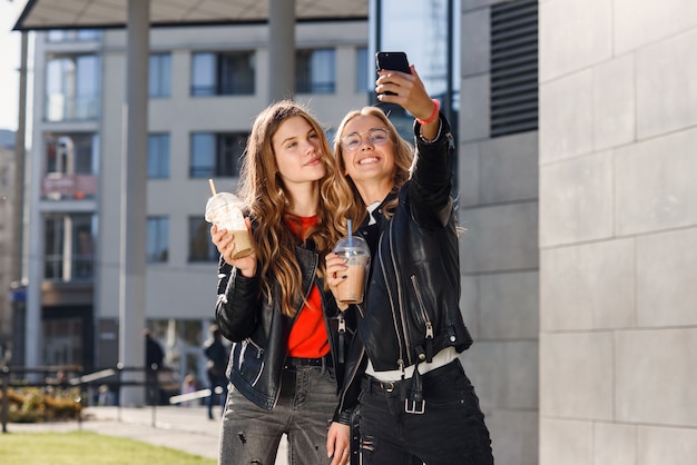 Zwei stilvolle attraktive Teenager-Mädchen mit Smoothie und Smartphone in der Nähe von Einkaufszentrum. Freizeit mit besten Freunden.