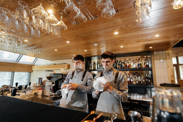 Zwei stilvolle Barkeeper in Masken und Uniformen während der Pandemie reiben Gläser, um zu glänzen Die Arbeit von Restaurants und Cafés während der Pandemie