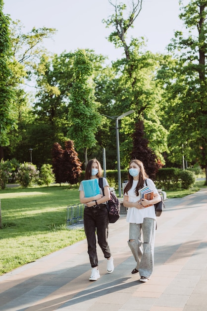 Zwei Studentinnen in medizinischen Schutzmasken gehen und unterhalten sich auf dem Campus. Fernstudium. Weicher selektiver Fokus.