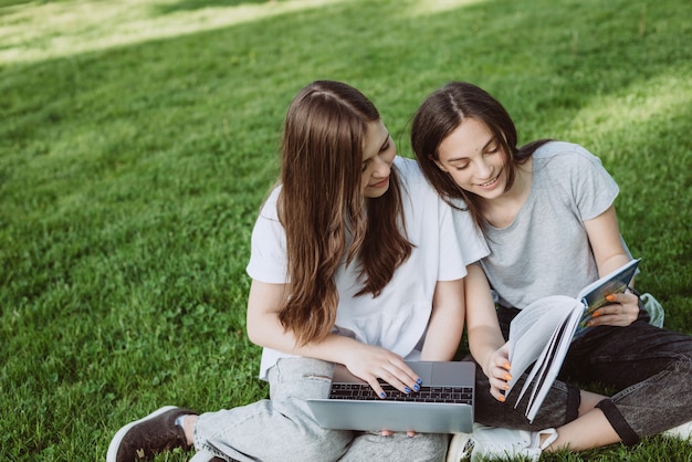 Zwei Studentinnen sitzen mit Büchern und Laptops im Park auf der Wiese, lernen und bereiten sich auf Prüfungen vor. Fernstudium. Weicher selektiver Fokus.