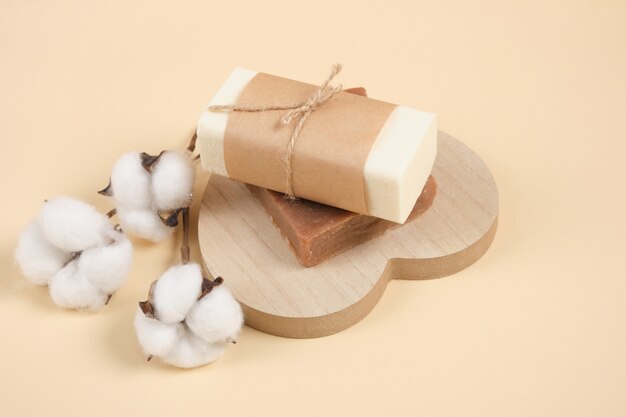 Zwei Stück hausgemachte handgemachte Seife auf einem Holzständer in Form eines Herzens und einer Baumwollblume, beigefarbener Hintergrund, umweltfreundliche Körperpflege