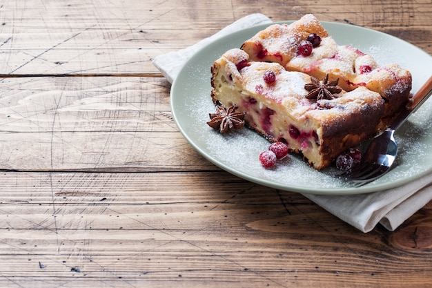 Zwei Stücke Hüttenkäse-Tortenkasserolle mit Moosbeerbeeren und Gewürzzimt und -anis auf einer Platte. Holzuntergrund.