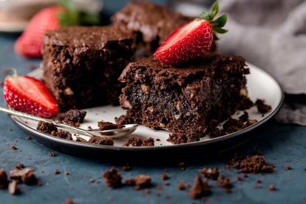 Zwei Stücke Schokoladenbrownie, Nahaufnahme, auf Untertasse mit Erdbeerscheiben, Krümel. Dunkelblauer Hintergrund