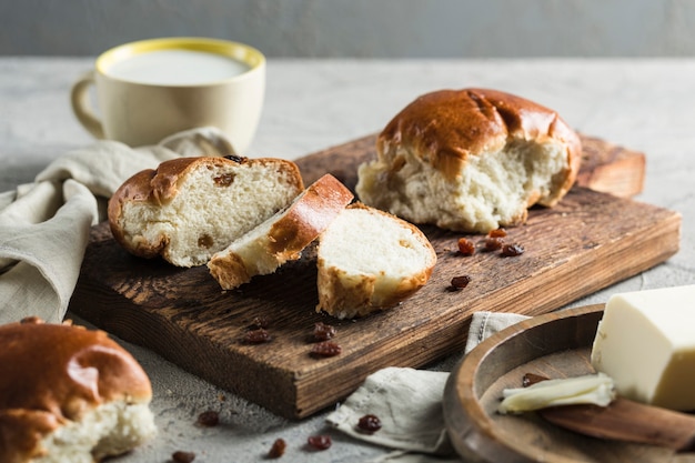 Zwei süße Brötchen mit Rosinen in Stücke geschnitten auf einem Holzbrett mit Butter und einer Tasse Milch