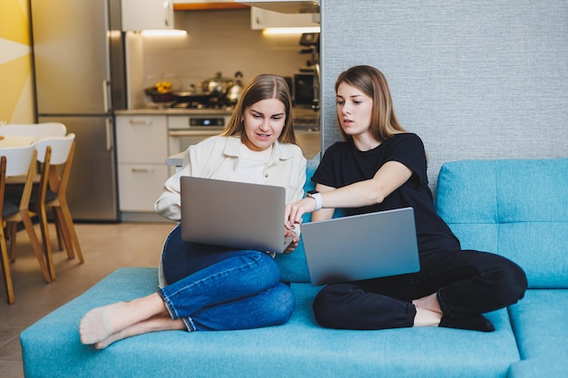 Zwei süße Frauen chatten mit ihren Freunden per Videoanruf über einen Laptop im Wohnzimmer. Freundliche Freundschaftszeit zusammen. Freundinnen sitzen auf der Couch und sehen sich einen Film an