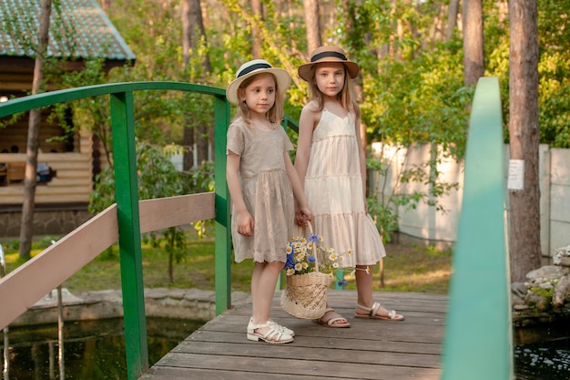 Zwei süße freundliche Tween-Schwestern in Sommerkleidern und Weidenhüten, die einen Korb mit Wildblumen halten, während sie entlang einer Holzbrücke über einen kleinen Teich im Hof des Landhauses gehen