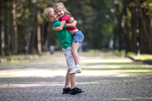 Zwei süße junge lustige lächelnde Kinder, Mädchen und Junge, Bruder, der Schwester in seinen Armen hält, Spaß auf verschwommenen hellen sonnigen Parkgassengrünbäumen hat. Liebevolles Geschwisterbeziehungskonzept.