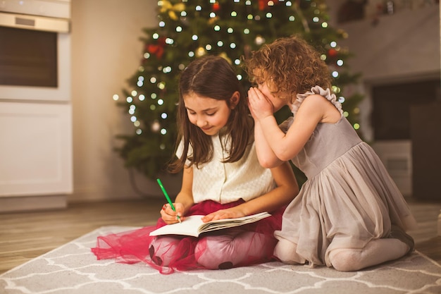 Zwei süße kleine Freundinnen schreiben zu Weihnachten in einem gemütlichen Wohnzimmer einen Brief an den Weihnachtsmann in der Nähe eines Tannenbaums