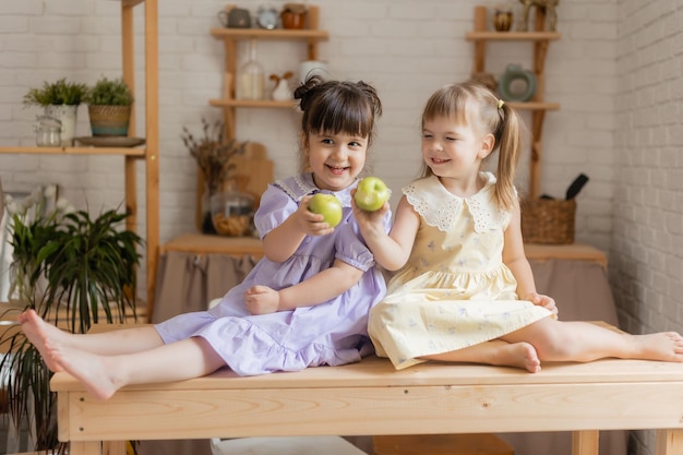 Zwei süße kleine lustige Mädchen in Kleidern essen grüne Äpfel in der Küche. Platz für Text, Banner