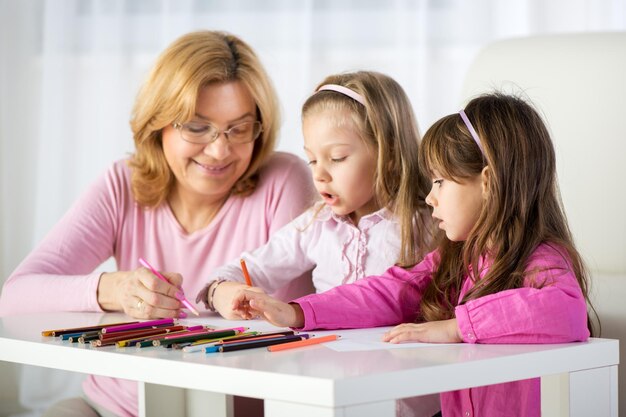 Zwei süße kleine Mädchen zeichnen zu Hause mit Großmutter mit Buntstiften.