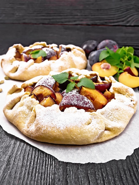 Zwei süße Kuchen mit Pflaume, Zucker und Kardamom auf Pergament, Minzzweige auf Holzbretthintergrund