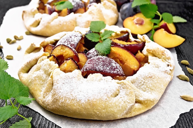 Zwei süße Kuchen mit Pflaumen, Zucker und Kardamom auf Papier, Zweige grüner Minze auf dunklem Holzbretthintergrund