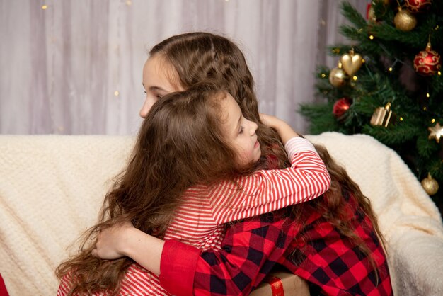 Zwei süße lächelnde Mädchen sitzen neben dem Weihnachtsbaum, haben einen Fächer und beschenken sich gegenseitig zum neuen Jahr