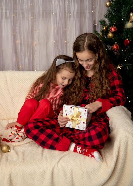 Zwei süße lächelnde Mädchen sitzen neben dem Weihnachtsbaum und beschenken sich gegenseitig mit Neujahrs- und Weihnachtsgeschenken