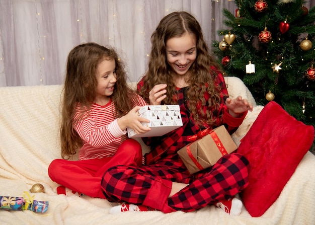 Zwei süße lächelnde Mädchen sitzen neben dem Weihnachtsbaum und beschenken sich gegenseitig mit Neujahrsweihnachten