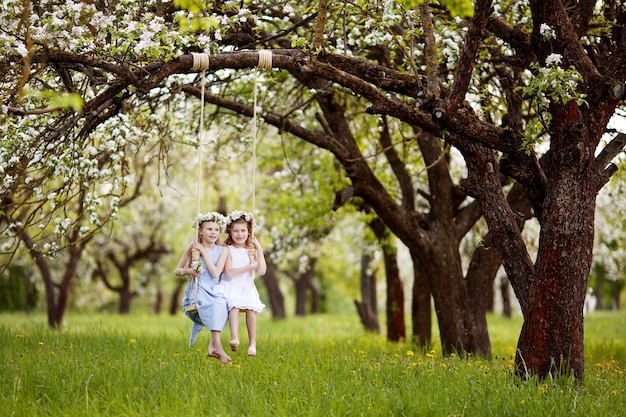 Zwei süße Mädchen, die Spaß auf einer Schaukel im blühenden alten Apfelbaumgarten haben. Sonniger Tag. Frühlingsaktivitäten im Freien für Kinder