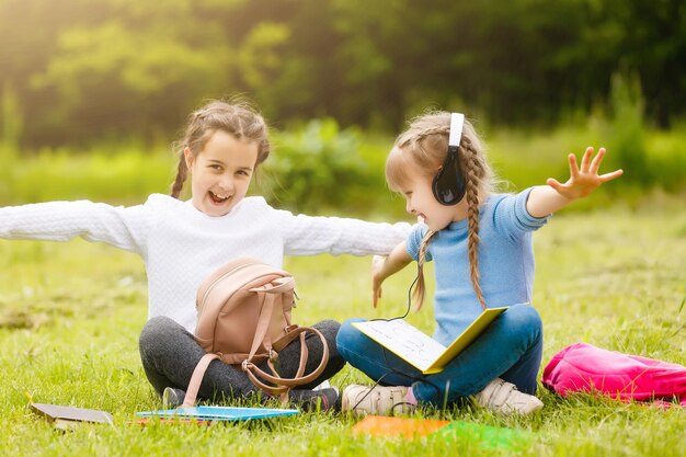 zwei süße multikulturelle Schulmädchen, die auf dem Rasen unter einem Baum sitzen und zusammen ein Buch lesen