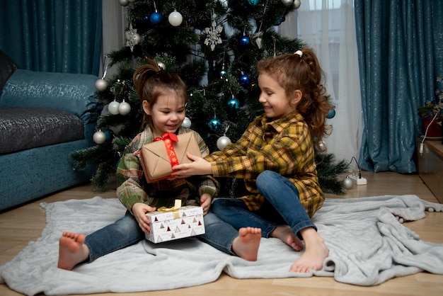 Zwei süße schöne Mädchen sitzen neben dem Weihnachtsbaum mit Geschenken Neujahr Weihnachten