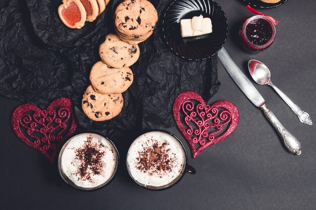 Zwei Tasse Kaffees, Cappuccino mit Schokoladenplätzchen und Kekse nähern sich roten Herzen auf schwarzer Tabelle