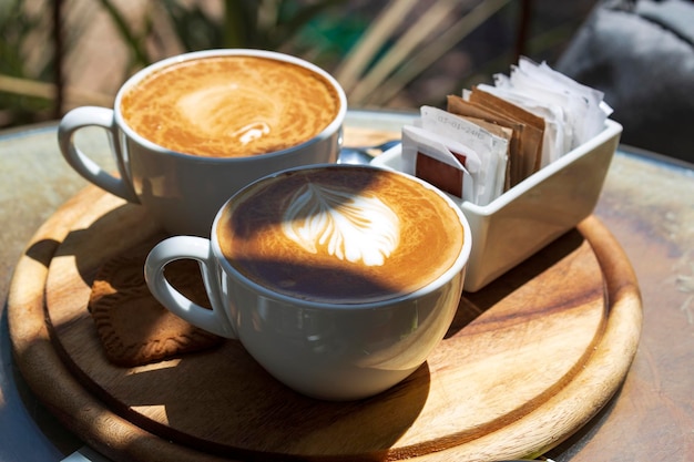 Zwei Tassen Cappuccino mit Latte Art auf Holztisch am Morgen bei direkter Sonneneinstrahlung Selektiver Fokus Inspirierendes Frühstück am frühen Morgen mit Kaffee Espresso-Latte-Kaffeetassen auf Holztablett