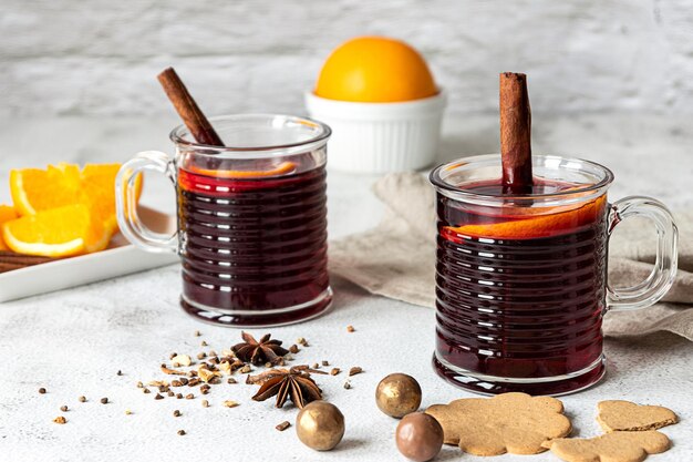 Zwei Tassen heißer Glühwein mit Zimtstangen, Gewürzen, Keksen und Orange. Rückwand aus grauem Beton