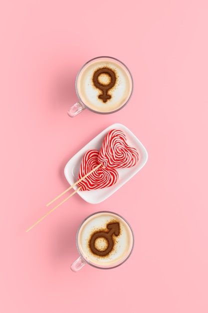 Zwei tassen kaffee mit symbolen von venus und mars auf schlagsahne-milchschaum und ein paar lutscher in herzform. pastellrosa hintergrund. konzept romantisches frühstück am valentinstag. flach liegen