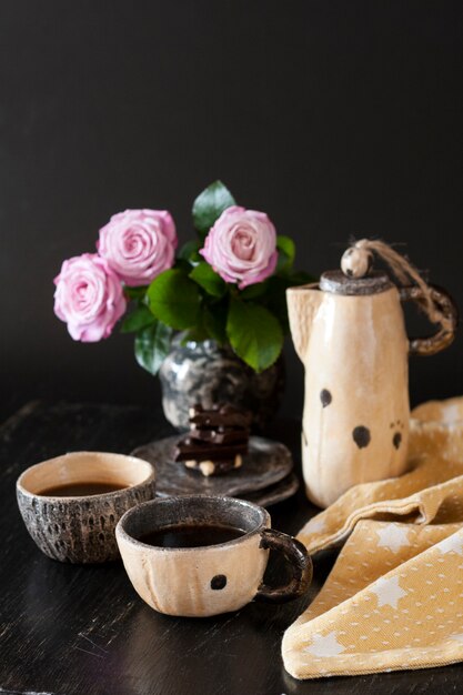 Zwei Tassen schwarzen Kaffee, schwarze Schokolade, eine gelbe Teekanne und eine Vase mit rosa Rosen auf dunkler Oberfläche