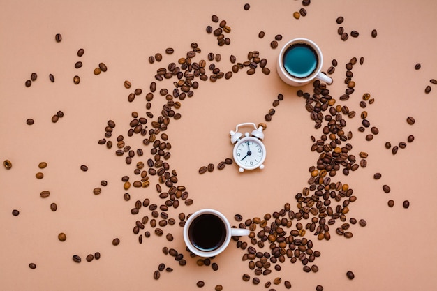 Zwei Tassen schwarzer Kaffee im Kreis aus Kaffeebohnen und ein weißer Wecker auf beigem Hintergrund. Zeit, Kaffeekonzept zu trinken. Ansicht von oben