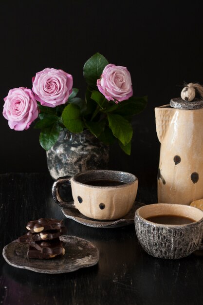 Zwei Tassen schwarzer Kaffee, schwarze Schokolade, eine gelbe Teekanne und eine Vase mit rosa Rosen