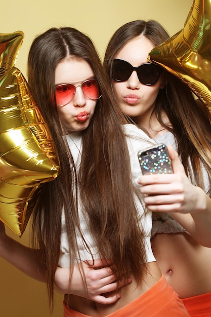 Zwei Teenager-Freundinnen mit goldenen Ballons machen Selfie auf einem Telefon Gelber Hintergrund