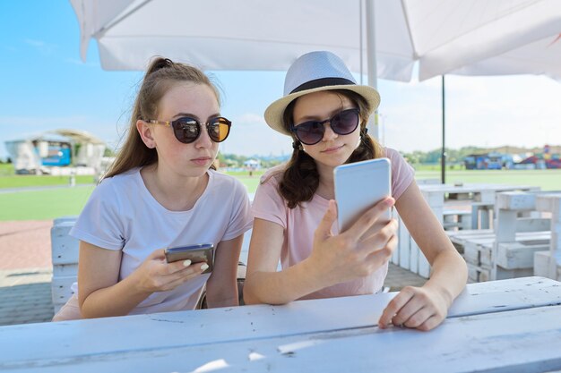 Zwei teenager-mädchen mit smartphones sitzen und sprechen im sommercafé im freien. jugend, teenager, freundschaft, kommunikation, menschenkonzept