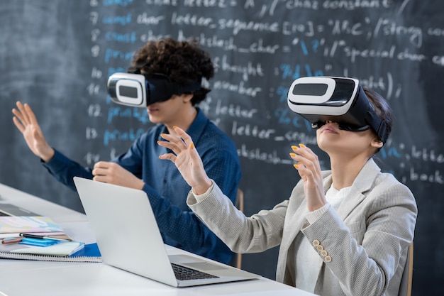 Zwei Teenager-Studenten in VR-Headsets sitzen am Schreibtisch an der Tafel und nehmen an einer Präsentation oder einem Seminar im Klassenzimmer im Unterricht teil