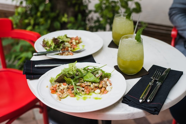 Zwei Teller mit frischem Salat mit Rucola, Mozzarella, Gemüse, Tomaten und Hühnchen werden zum Mittagessen in einem kleinen Restaurant im Freien mit Minzlimonade serviert