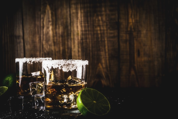 Zwei Tequila-Schnapsgläser auf dunklem Hintergrund, mit Eiswürfeln, Salz und Kalken