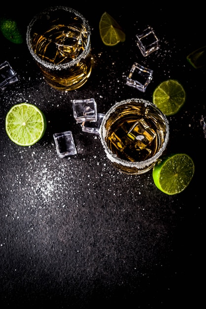 Zwei Tequila-Schnapsgläser auf dunklem Tisch, mit Eiswürfeln, Salz und Limetten