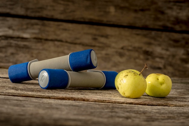 Zwei Trainingshanteln und drei frische reife Äpfel, die auf einem strukturierten rustikalen Holzschreibtisch liegen Konzept von Bewegung und gesundem Lebensstil