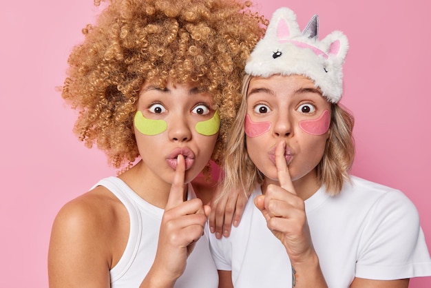 Zwei überraschte junge Frauen mit Augenflecken, um Falten zu entfernen, genießen Hautpflegebehandlungen, die eng beieinander stehen, und machen stille Gesten, die das Geheimnis der Schönheit teilen, isoliert auf rosafarbenem Hintergrund