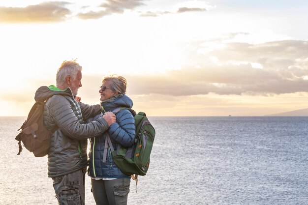 Zwei unbeschwerte ältere Menschen mit Rucksäcken schauen sich in die Augen und genießen das Wintermeer. Ein schönes älteres Paar, das auf der Klippe mit Horizont über dem Wasser und Sonnenuntergang steht