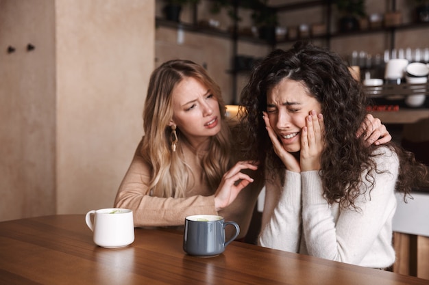 Zwei verärgerte junge Freundinnen, die am Cafétisch sitzen, Kaffee trinken und sich gegenseitig aufmuntern