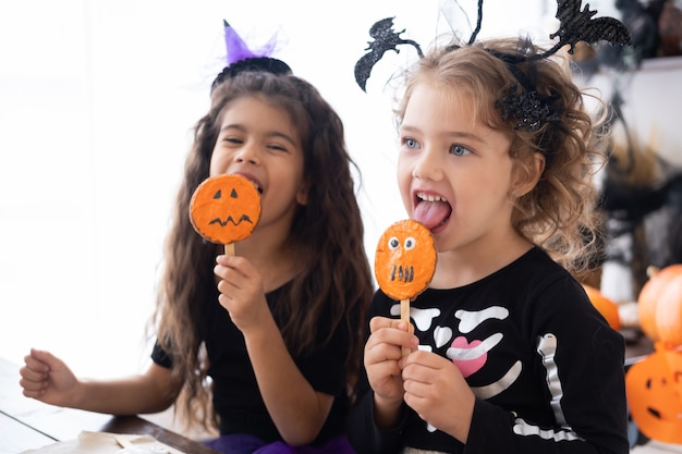 Foto zwei verschiedene kindermädchen im kostüm der hexe, die spaß in der küche haben, kekse essen, halloween feiern.