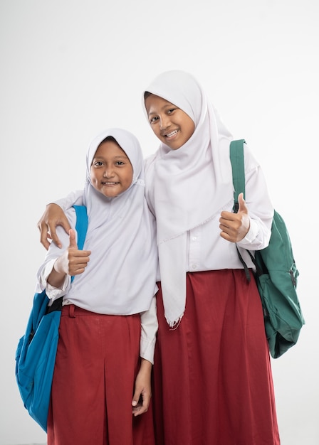 Zwei verschleierte Mädchen in Grundschuluniformen mit Daumen nach oben, während sie einen Rucksack und ein Buch tragen