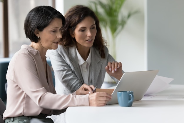Zwei vielfältige Geschäftsfrauen, Millennial und reife Kollegen, sprechen, schauen auf den Laptop-Bildschirm, überprüfen die App, diskutieren über eine Online-Präsentation, arbeiten an einem gemeinsamen Projekt, teilen Ideen während der Teamarbeit.