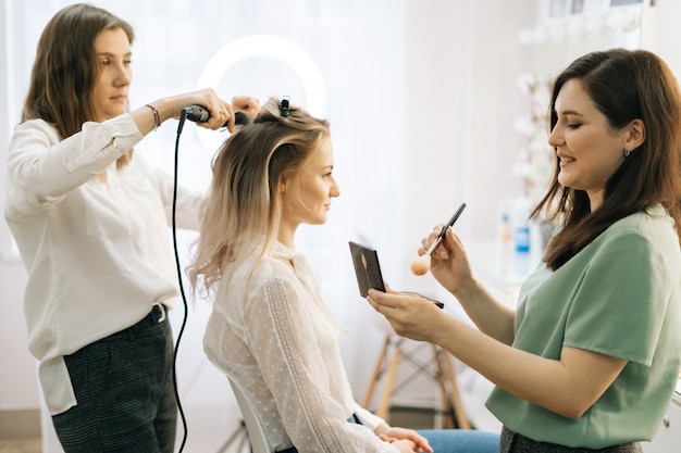 Zwei Visagisten tragen der Braut in der Umkleidekabine Haare und Make-up auf