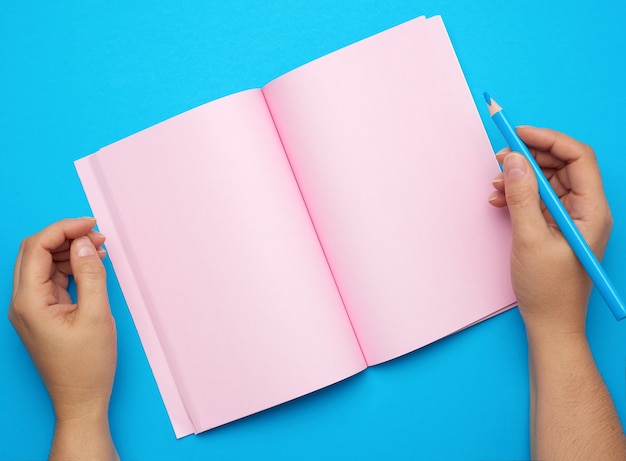 Zwei weibliche Hände, die offenen Notizblock mit leeren rosa Blättern auf einem blauen Hintergrund halten